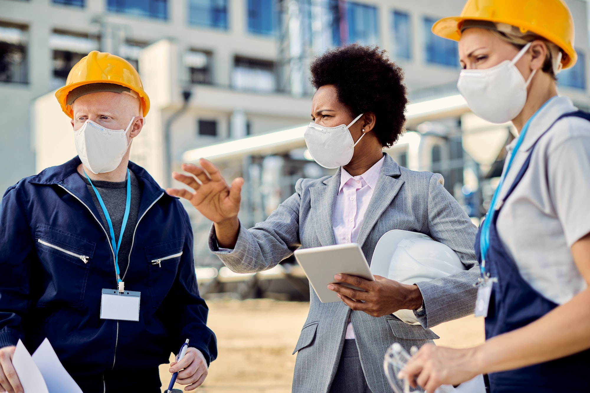 Black female architect and civil engineers with protective face masks talking at construction site.